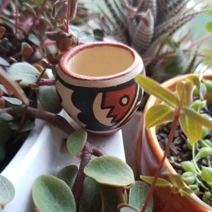 May include: A small, hand-painted ceramic pot with a black, white and red geometric design. The pot is sitting on a white surface with green plants in the background.