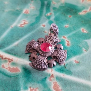 May include: A silver pendant with a red gemstone in the center, surrounded by intricate silver designs. The pendant is shaped like a cross.