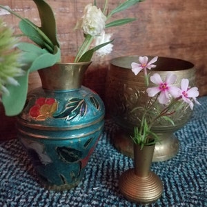 May include: Three brass vases with floral arrangements. The largest vase is engraved with a floral pattern and holds pink flowers. The medium vase is painted with green, blue, and red floral designs and holds white flowers. The smallest vase is plain and holds pink flowers.