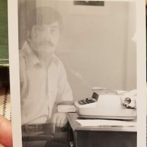 May include: Black and white photograph of a person sitting at a desk with a typewriter. The person is wearing a striped shirt and has a mustache. The typewriter is white and has black keys.
