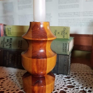 May include: A wooden candle holder with a natural wood finish. The holder has a wide base and a narrow top, with a cylindrical shape in the middle. The holder is made of two different types of wood, creating a striped pattern.