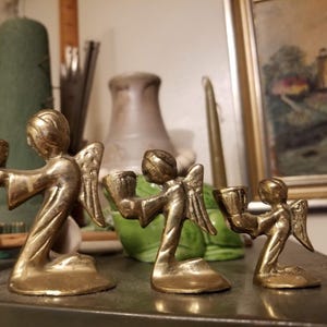 Peut inclure: Trois bougeoirs en laiton en forme d'anges de tailles différentes. Chaque ange est agenouillé et tient un bougeoir en forme de coupe. Les anges ont des ailes détaillées et une finition dorée. L'arrière-plan est flou.