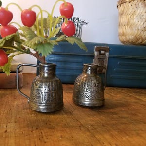 May include: Two small silver pitchers with intricate designs, sitting on a wooden surface. A bouquet of red plastic berries with green leaves is in the background.