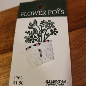 May include: A white button with a flower pot design, featuring a textured pot and a floral arrangement. The card reads "FLOWER POTS" and includes the number "1762". The button is from Blumenthal Lansing Crafts.