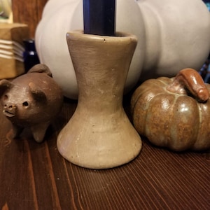 May include: A brown ceramic candle holder with a wide base and a narrow top. The holder is sitting on a dark wooden surface with a small brown ceramic pig and a brown ceramic pumpkin beside it.