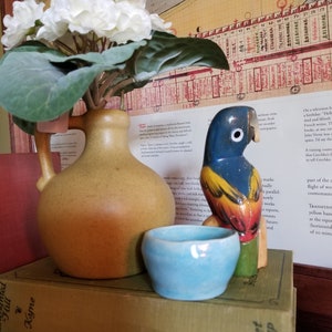May include: A colorful wooden parrot figurine with blue, yellow, and red paint sits on a stack of books. The books are titled "Hill Led Kyne" and "Grosset & Dunlap". A small blue ceramic bowl and a brown ceramic vase with white flowers are also on the books.