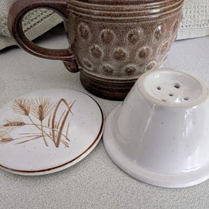 May include: A ceramic tea cup and a matching tea pot with a lid. The cup is brown with a textured pattern. The tea pot is white with a flared shape and small holes. The lid is white with a wheat design.