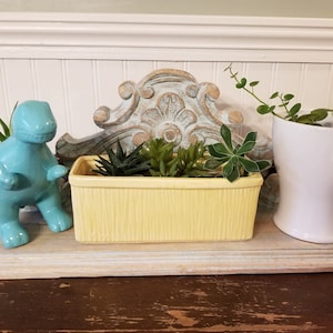 May include: A decorative shelf arrangement featuring a light blue ceramic dinosaur figurine, a yellow rectangular planter with succulents, and a white ceramic pot with greenery. A distressed wooden decorative element is behind the planter.