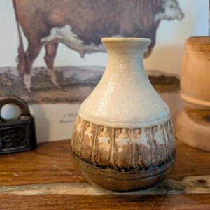 May include: A small, handcrafted ceramic vase with a cream-colored upper portion and a textured, brown lower section. The vase has a rounded body and a narrow neck. The background includes a framed bull illustration and wooden objects.