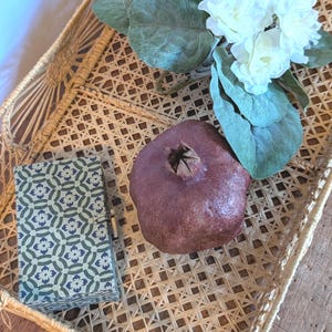 May include: A rectangular woven rattan tray with a lattice pattern, containing a small rectangular box with a blue and green floral geometric print, a dark purple pomegranate-shaped object, and a cluster of white artificial flowers with green leaves.