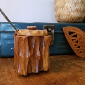 May include: A wooden jar with a lid and a spoon. The jar is carved with a wood grain pattern and has a natural wood finish. The lid is also made of wood and has a smooth surface. The spoon is made of metal and has a simple design.
