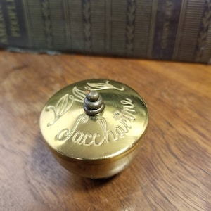 May include: A gold-colored metal trinket box with a lid. The lid has a decorative design and the words "Saccharine" engraved on it.