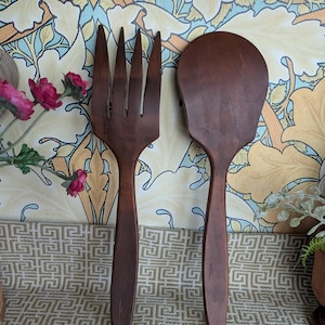 May include: A dark brown wooden fork and spoon set. The fork has four tines, and the spoon has a wide, flat head. The set is displayed against a patterned background with floral and geometric designs. A decorative kitchen utensil set.
