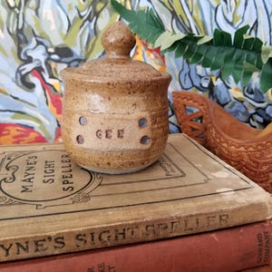 Puede incluir: Un pequeño tarro de cerámica marrón con tapa. El tarro tiene un esmalte moteado y una forma redonda. La tapa tiene un pomo en la parte superior. El tarro tiene la palabra "GEE" escrita en la parte delantera. El tarro está sentado encima de dos libros.