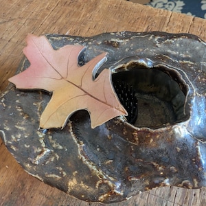 May include: A handcrafted, dark brown ceramic bowl with a textured surface. A decorative, multi-toned leaf rests on the bowl's edge. The bowl has a central opening, suggesting it could be used as a decorative vase or a unique home decor item.