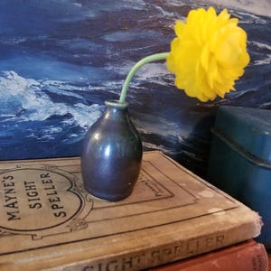 May include: A single yellow flower in a small, dark blue and green ceramic vase. The vase is sitting on top of two vintage books, one with the title "Mayne's Sight Speller".