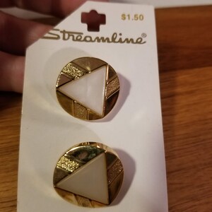 May include: Two gold-toned earrings with a white triangle design, set on a white card. The earrings have a circular shape with a textured gold border and a white triangle in the center. The card reads "Streamline" and "WHITE".