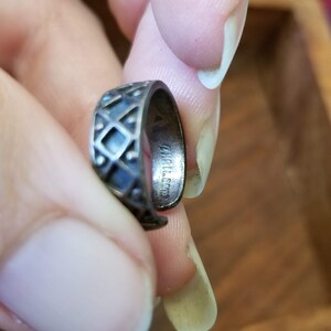 May include: A silver ring with a geometric diamond pattern. The ring is held between fingers, and the inside of the ring has the text "THAILAND". The ring is a dark silver color.