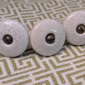 May include: Three round ceramic knobs with a glossy off-white finish and a visible dark crackle pattern. Each knob features a centered, dark metallic screw head with a flat-head slot, providing a functional attachment point for furniture hardware.