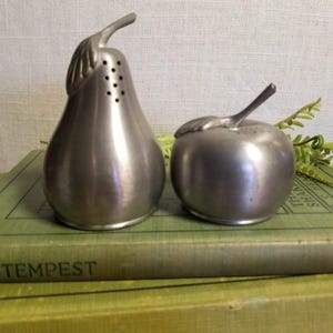 May include: A set of two silver salt and pepper shakers shaped like a pear and an apple. The shakers are sitting on top of two green books.