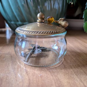 May include: A small, clear glass jar with a brass-colored lid and rim, containing several antique keys. The lid is topped with a decorative finial and a small, amber-colored bead. The jar sits on a wooden surface.