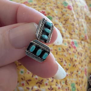 May include: A pair of rectangular silver earrings with three oval turquoise stones. The earrings have a braided border and are held between fingers against a yellow floral background. The earrings are a Southwestern style.