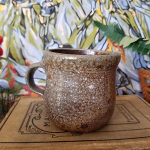 May include: A speckled brown and gray ceramic mug with a rounded body and a handle. The mug is sitting on top of a vintage book with the title "Mayne's Sight Spell".