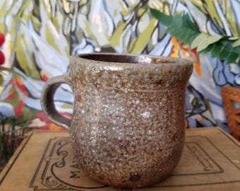 May include: A speckled brown and gray ceramic mug with a rounded body and a handle. The mug is sitting on top of a vintage book with the title "Mayne's Sight Spell".