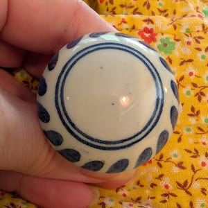 May include: A round, white ceramic knob with a blue floral design. The knob has a central blue circle and is surrounded by blue teardrop shapes. The knob is held against a yellow floral fabric.