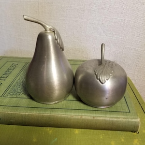 May include: A pair of pewter-colored salt and pepper shakers shaped like a pear and an apple. The pear shaker has a stem and leaf detail, while the apple shaker has a stem and leaf detail with small holes for dispensing. They are sitting on a green book.