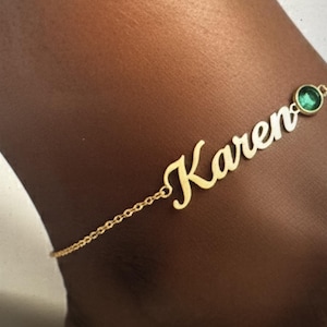 Può includere: Cavigliera dorata con il nome "Karen" in corsivo. Una piccola gemma verde rotonda è incastonata vicino alla chiusura. La delicata catena è attaccata al nome e alla gemma.
