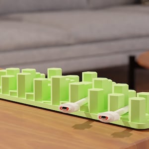 Puede incluir: Un organizador de cables verde claro con un diseño de paisaje urbano. El organizador tiene múltiples bloques rectangulares verticales y dos cables blancos con conectores rojos. El artículo está sobre una mesa de madera.
