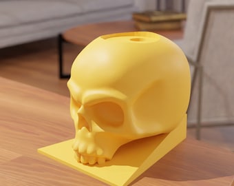 May include: A yellow skull-shaped planter with a small hole on top. The planter is sitting on a yellow base.