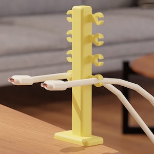May include: A yellow cable organizer with multiple clips to hold cords in place. The organizer is mounted on a wooden surface and has two white cables attached to it.