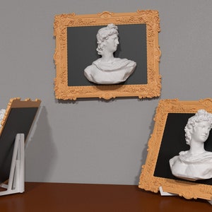 以下が含まれることがあります： 黒いマットの入った、金色の装飾が施されたフレームが3つあり、月桂樹の冠をかぶった人物の白い石膏の胸像が飾られています。フレームは木製のテーブルの上に置かれています。