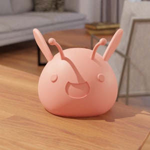 Pode incluir: Uma criatura rosa, de tipo cartoon, com duas antenas e um sorriso largo. A criatura é feita de um material suave e liso e tem uma forma arredondada.