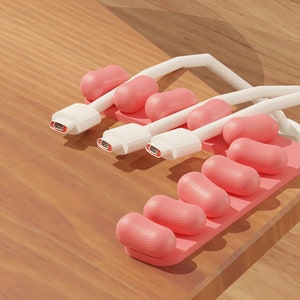 May include: A pink and white cable organiser with six slots for holding cables. The organiser is made of a soft, flexible material and has a unique design that resembles a row of jelly beans.