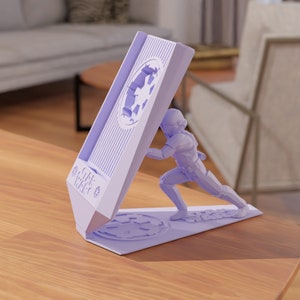 Puede incluir: Un soporte para teléfono de color morado impreso en 3D con un diseño de Star Wars. Una figura de Stormtrooper está representada empujando contra una pared con el símbolo de la Estrella de la Muerte. El soporte tiene las palabras "Star Wars" impresas en el lateral.