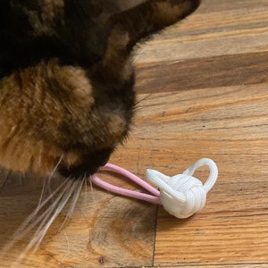 May include: A cat's face with brown and black fur is close to a white and pink rope toy. The toy is a small, decorative teapot shape made of knotted white rope, with a pink rope handle.