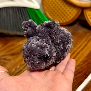 May include: A small, grey plush bunny with black eyes. The handmade stuffed animal is held in a person's hand. The bunny has floppy ears and a fluffy texture, suggesting it is made of soft yarn or fabric.