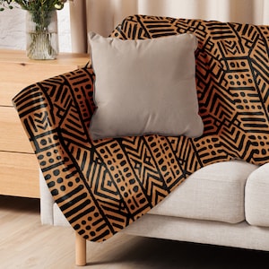 May include: A beige sofa with a tan throw blanket featuring a black geometric design. A plain tan cushion sits on the blanket. The sofa has wooden legs and is set against a neutral backdrop.