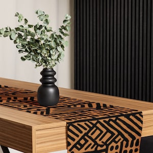 May include: A wooden table with a decorative runner featuring a black geometric pattern on a tan background. A black vase holds green eucalyptus branches. The table is set against a black slatted wall.