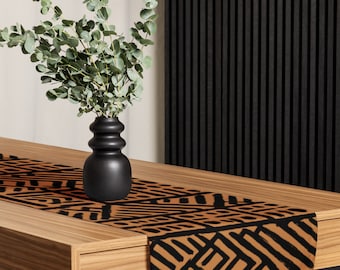 Può includere: Un tavolo in legno con un runner decorativo con un motivo geometrico nero su sfondo marrone. Un vaso nero contiene rami di eucalipto verdi. Il tavolo è appoggiato a una parete a doghe nere.