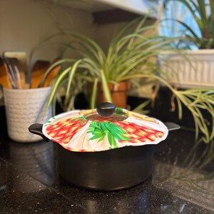 May include: A black cooking pot with a glass lid and a colourful pineapple-patterned cover. The cover features red, yellow, and green colours. The pot is on a black speckled countertop, with plants in the background.