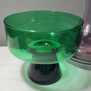May include: A green glass pedestal bowl with a dark green base. The bowl is clear and has a rounded shape.