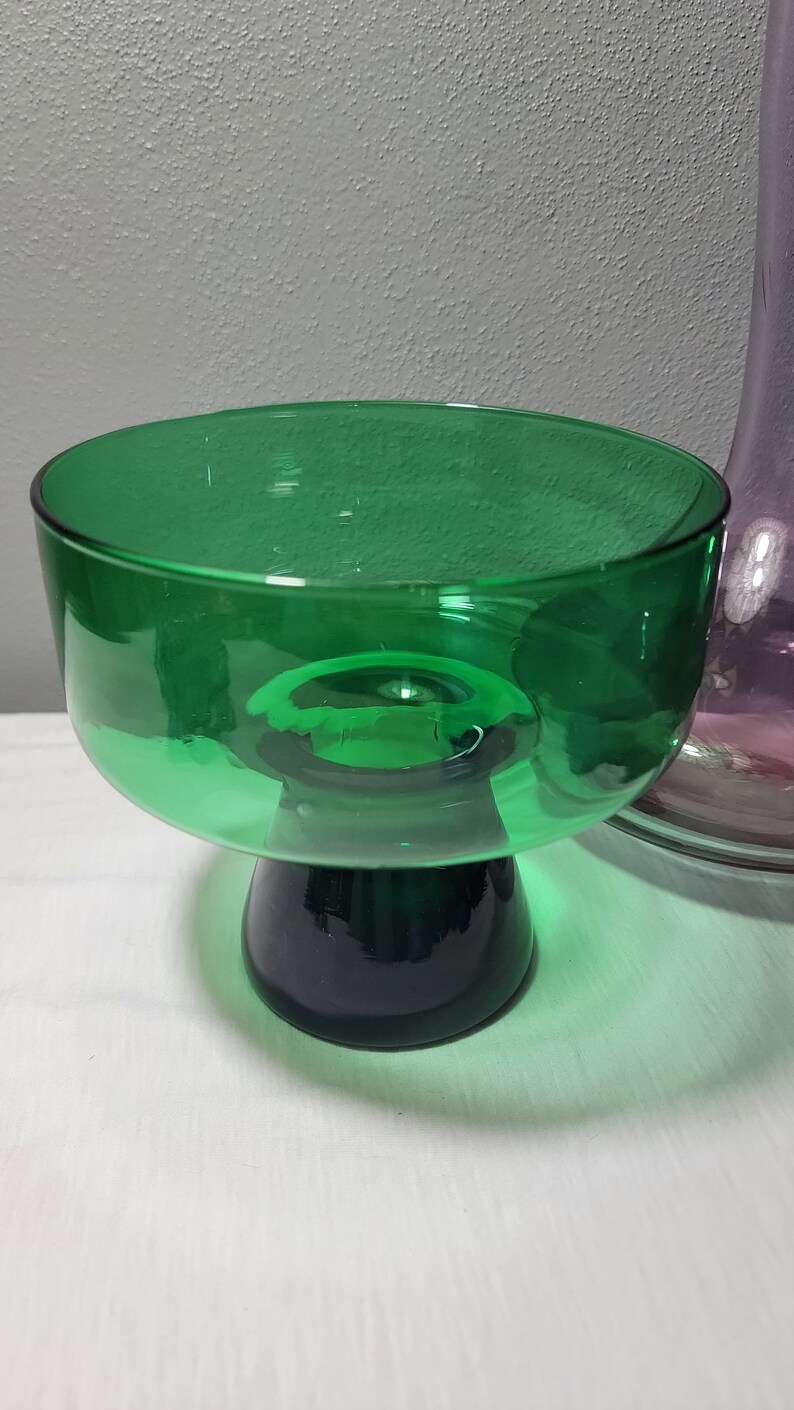 May include: A green glass pedestal bowl with a dark green base. The bowl is clear and has a rounded shape.