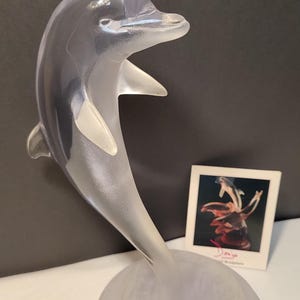 May include: A clear glass sculpture of a dolphin leaping out of the water. The dolphin is on a frosted glass base with a wave design. The sculpture is signed "Dalia Jo" on a small card.