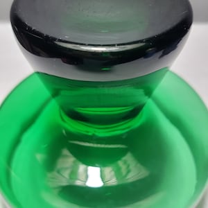May include: A green glass bottle stopper with a black top. The stopper has a round base and a raised, rounded top.