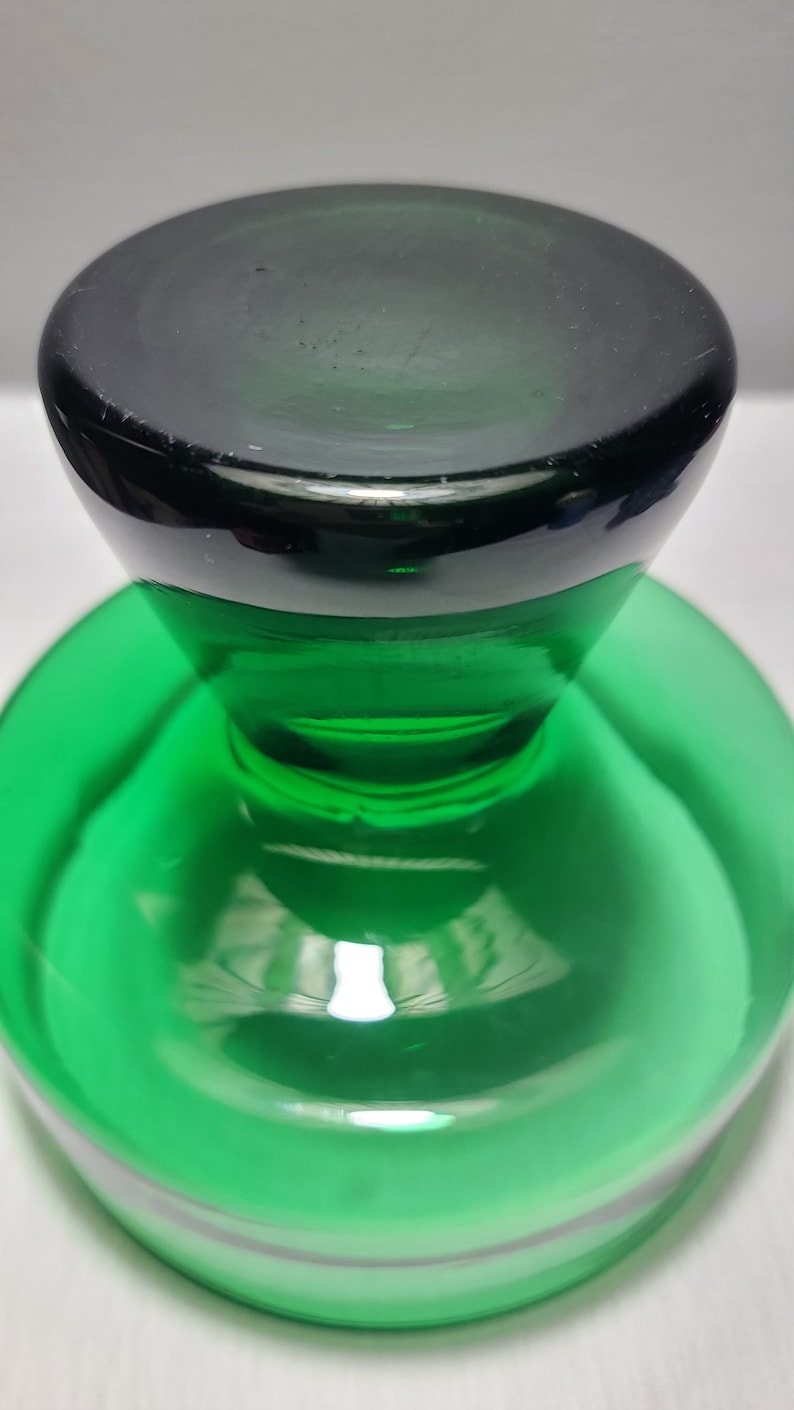 May include: A green glass bottle stopper with a black top. The stopper has a round base and a raised, rounded top.