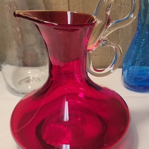 May include: A ruby red glass pitcher with a clear glass handle in the shape of a figure eight. The pitcher has a round body and a wide mouth.